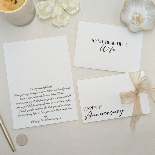 Luxury 1st wedding anniversary paper gift card featuring 'Happy First Anniversary' in black ink, finished with a hand-tied gold wired-edge organza ribbon. The card is shown open with a personalised printed message inside and a matching custom-printed envelope.
